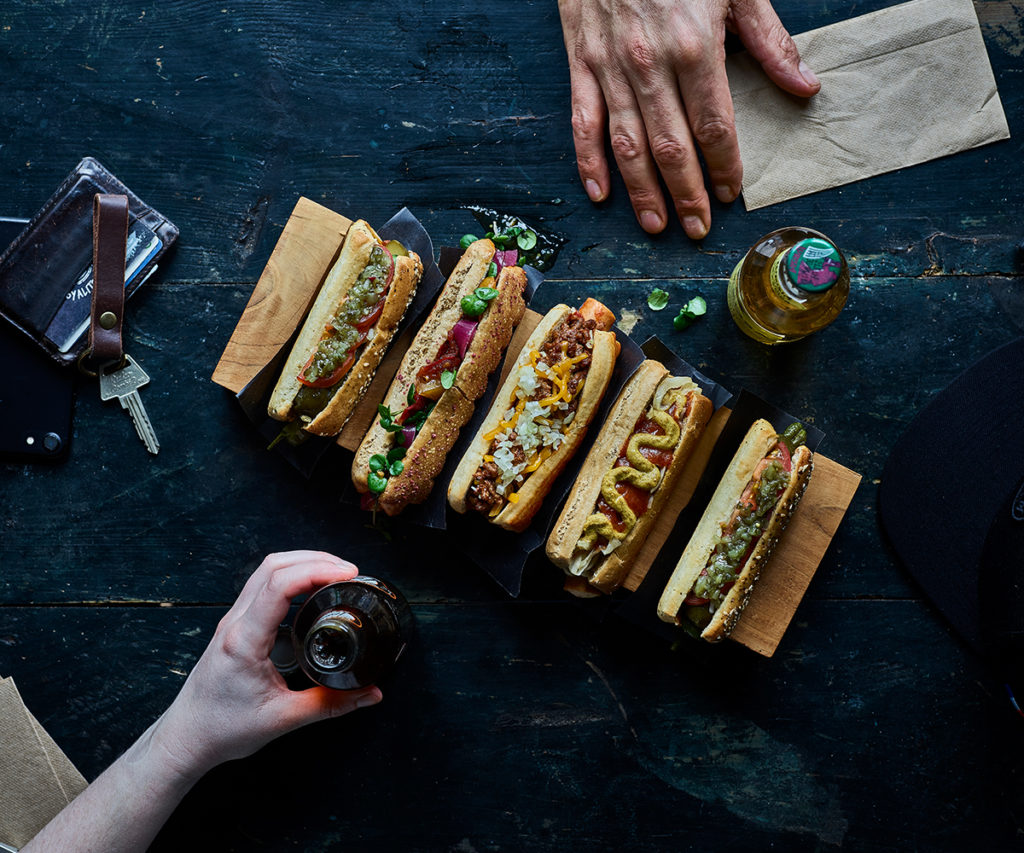 4 hot dogs with toppings, two hands in frame