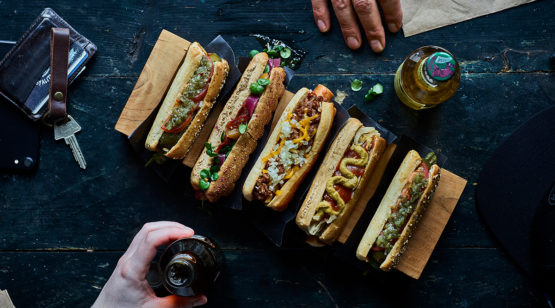 4 hot dogs with toppings, two hands in frame